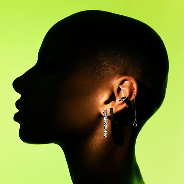 A close-up side profile of a person with a shaved head wearing multiple silver earrings against a bright lime green background. One earring is a long rugged bar, a smooth bar a square ear cuff and another is a dangling mini hexagon threader.