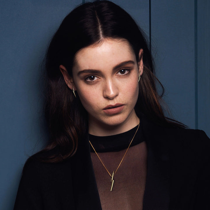 A close-up portrait of a woman with dark hair wearing Edge Only Skinny Lightning Bolt Pendant necklace and 3D Pointed Lightning Bolt Earrings 14ct Gold. Set against a dark blue paneled background.