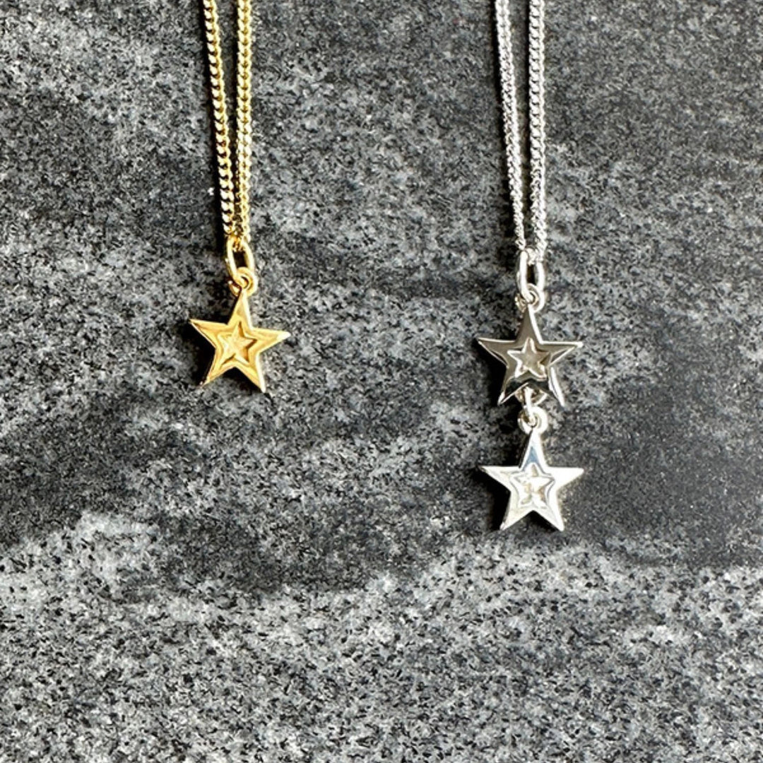 Two star pendant necklaces displayed on a textured gray stone surface. One necklace has a gold chain and a gold star pendant, while the other has a silver chain with two silver star pendants.