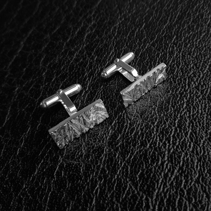 A pair of silver-toned rectangular cufflinks with textured, faceted edges, displayed on a black textured leather background. Product is Edge Only Rugged Cufflinks in Sterling Silver.